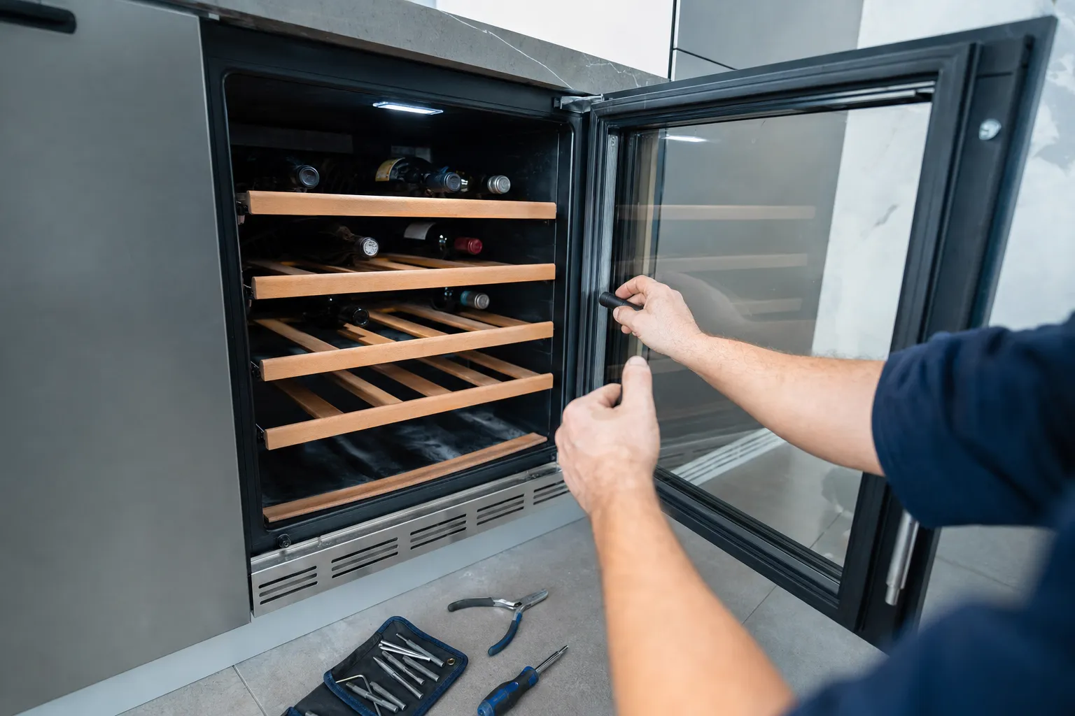 Glass door held open on an undercounter wine cooler with wooden racks, a tool roll and pliers on the floor