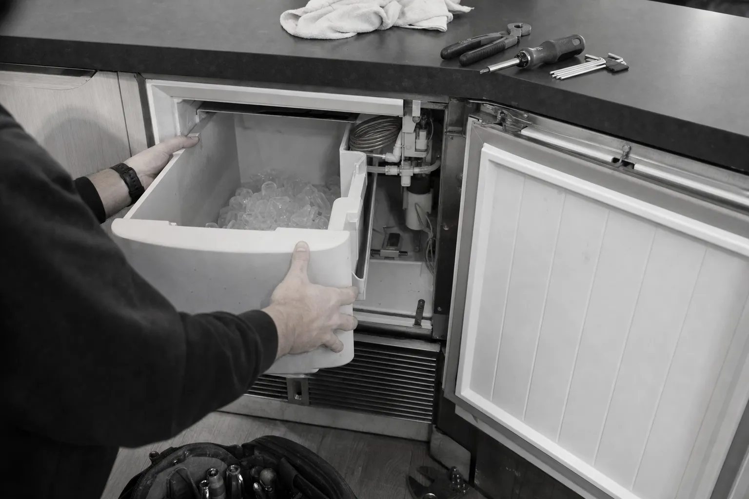 Black-and-white shot of an ice bin drawn from an undercounter ice maker, door panel off and water line exposed