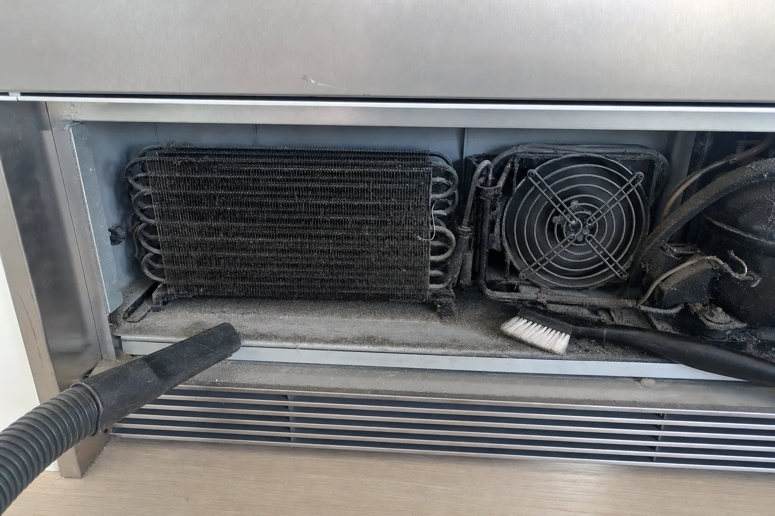 Dusty condenser coil and fan guard exposed under a stainless unit, a vacuum nozzle and soft brush at the coil, louvred grille below